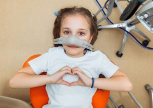 Young girl making heart shape with hands while wearing nitrous oxide mask